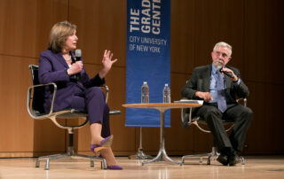 Two people sit in chairs on a stage, a small table between them and a blue banner behind them (with Graduate Center logo). Speaker Emerita Nancy Pelosi has raised the microphone and is speaking and gesticulating; Prof. Paul Krugman is listening attentively. On the stage of the Graduate Center, CUNY